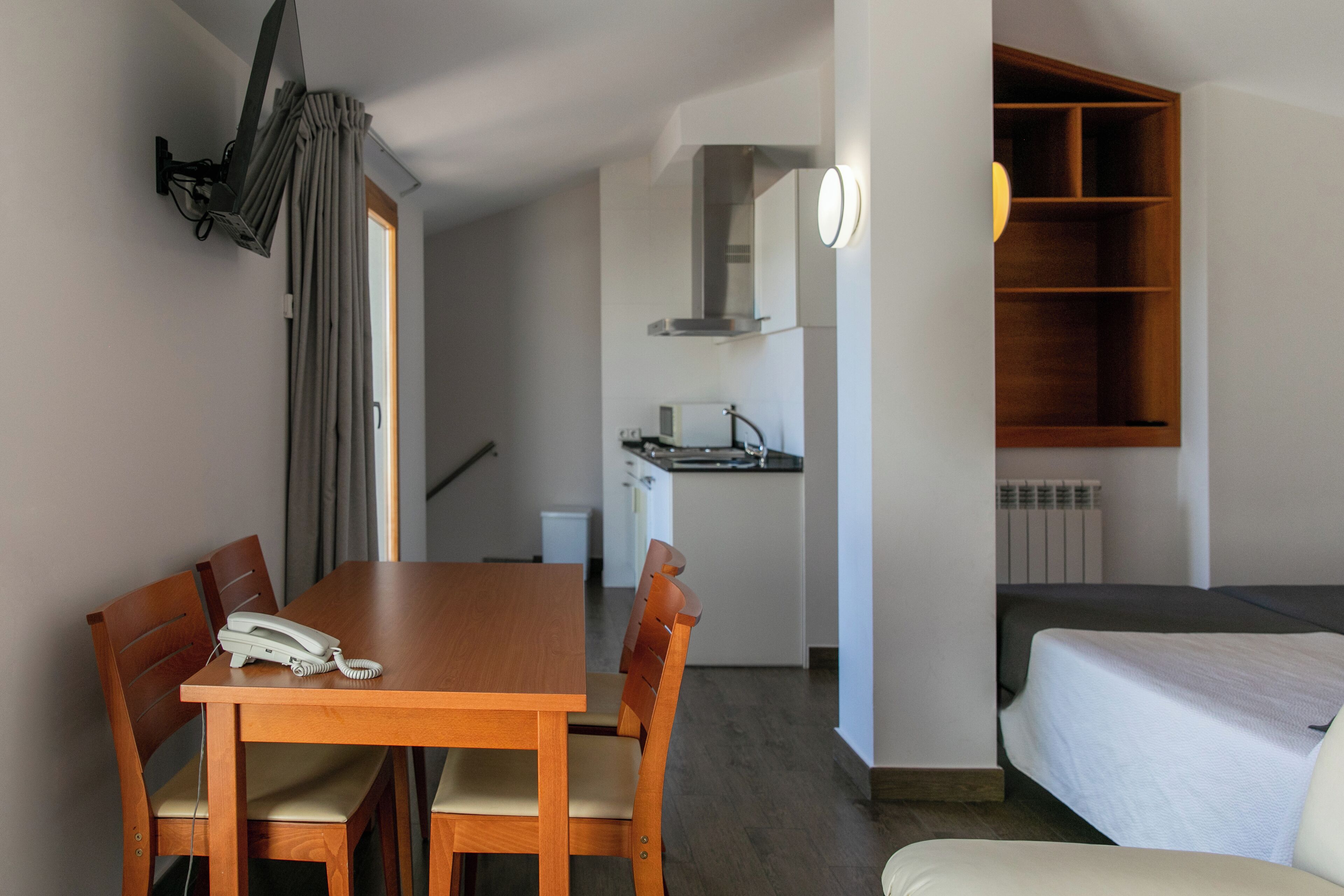 A compact hotel room with a wooden dining table, four chairs, a single bed, a small kitchenette, and a built-in shelf.
