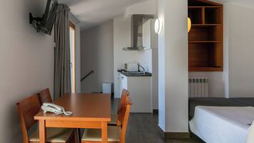 A compact hotel room with a wooden dining table, four chairs, a single bed, a small kitchenette, and a built-in shelf.