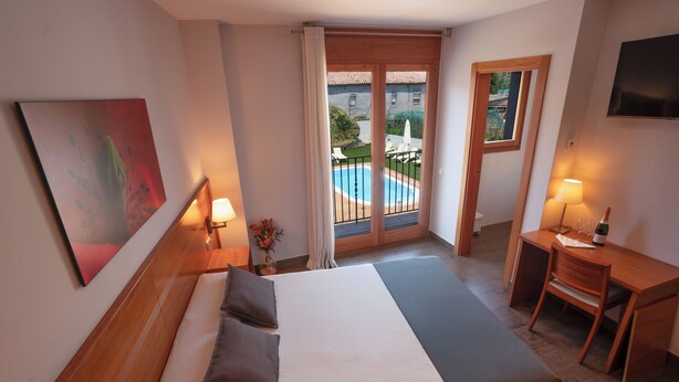 Habitación de hotel con cama, escritorio, silla, lámpara, un cuadro y vista a una piscina y un edificio a través de una puerta de vidrio.