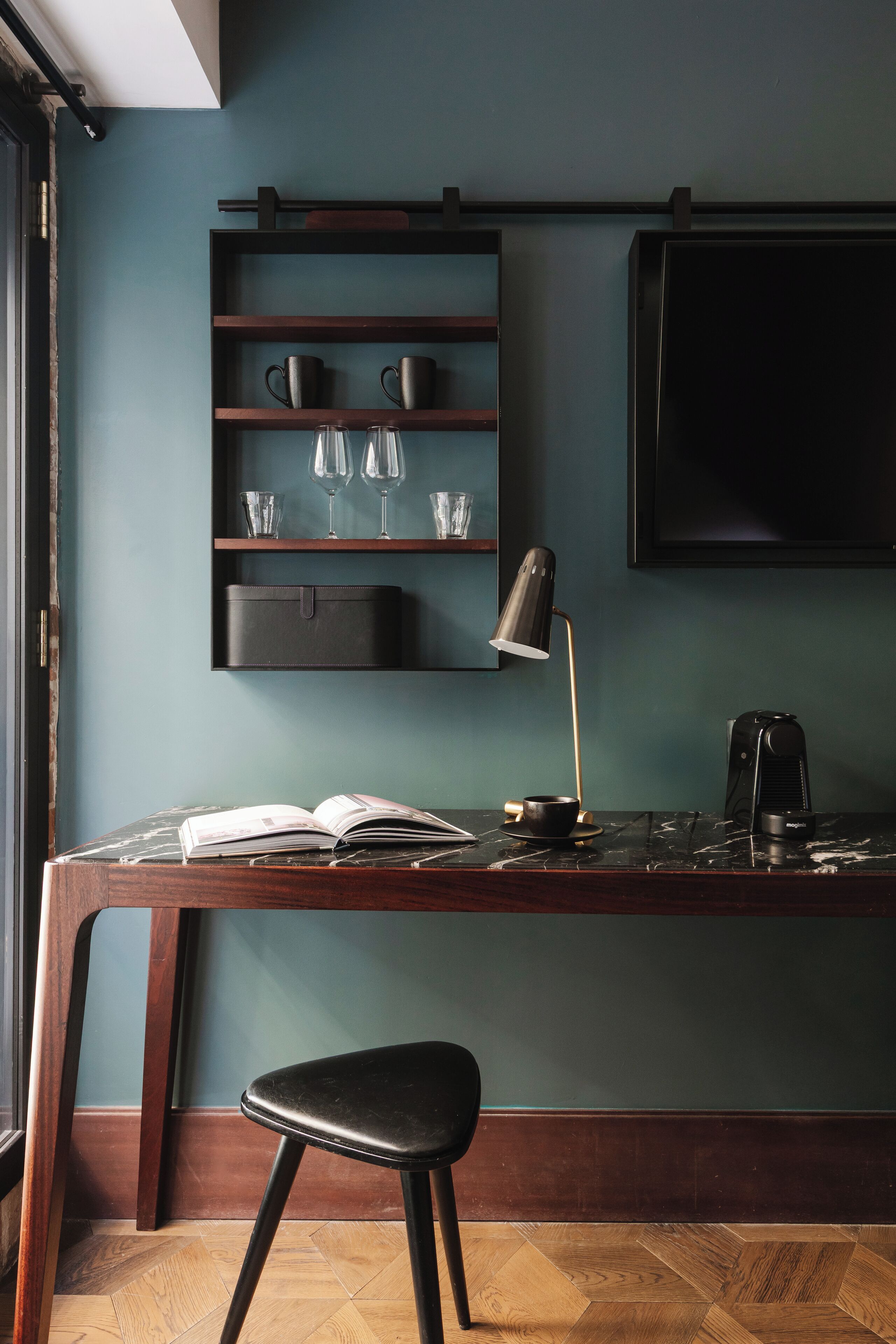A desk with a book, a cup, and a coffee maker. There is a shelf above the desk holding cups and glasses. A chair is in front of the desk.