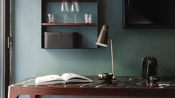 A desk with a book, a cup, and a coffee maker. There is a shelf above the desk holding cups and glasses. A chair is in front of the desk.