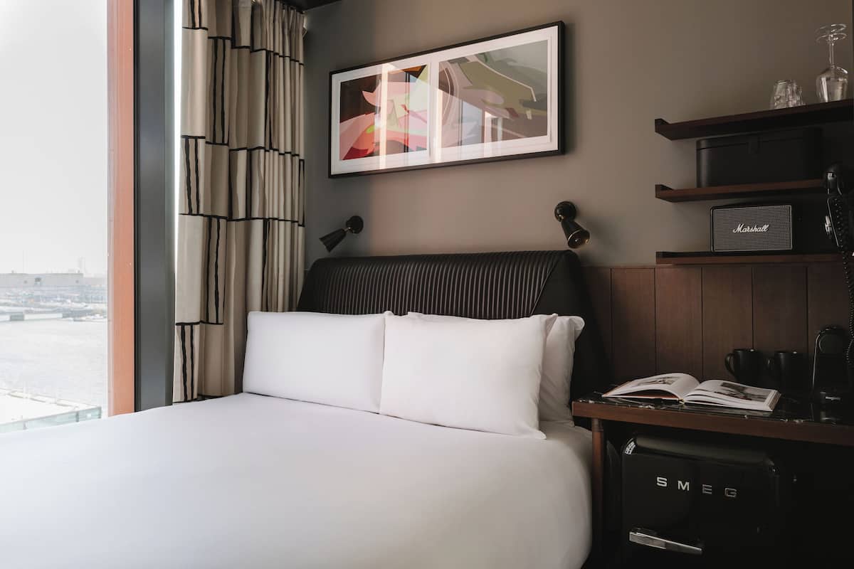Una habitación de hotel con una cama, una obra de arte enmarcada en la pared, un escritorio con un teléfono y un libro, y vistas a la ciudad por la ventana.