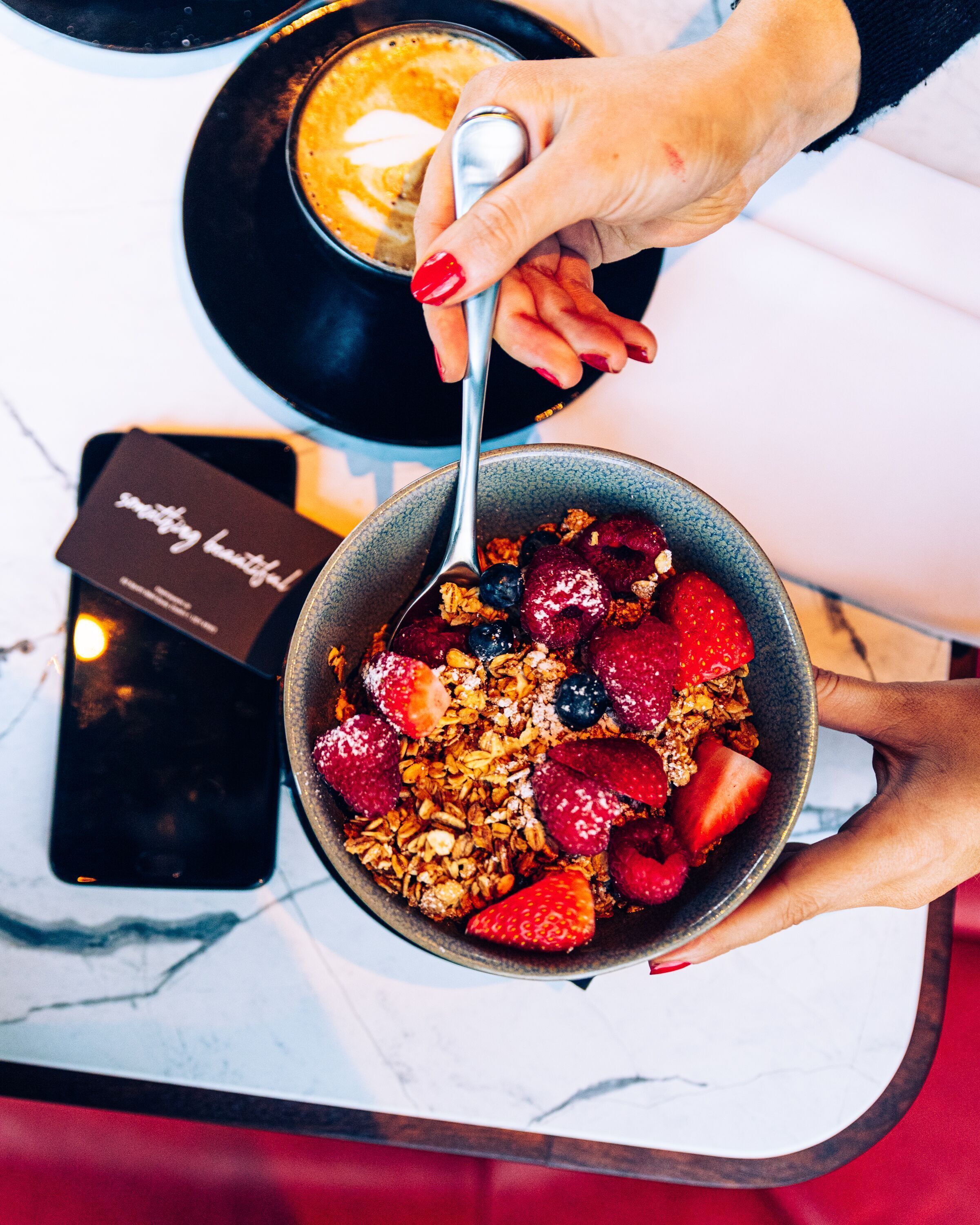 A bowl of granola with berries and a spoon, a smartphone, and a coffee cup on a table.
