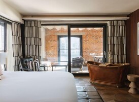 Una camera d'albergo con un letto grande, una vasca da bagno, un'area pranzo con tavolo e sedie, e vista su un edificio in mattoni attraverso la porta vetrata.
