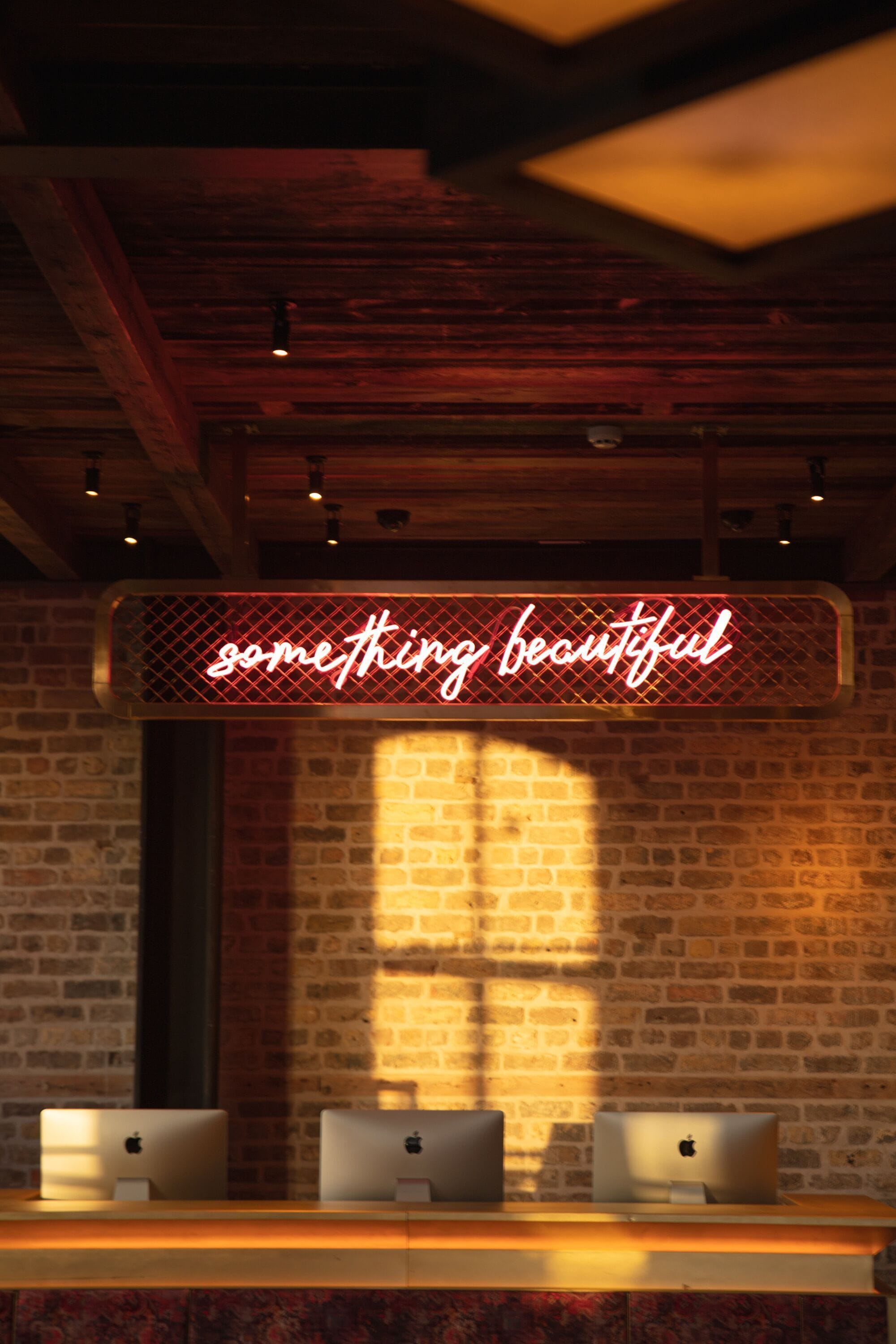 A room with a brick wall, wooden ceiling beams, and a neon sign that reads "something beautiful." There are three Apple iMac computers on a wooden desk.
