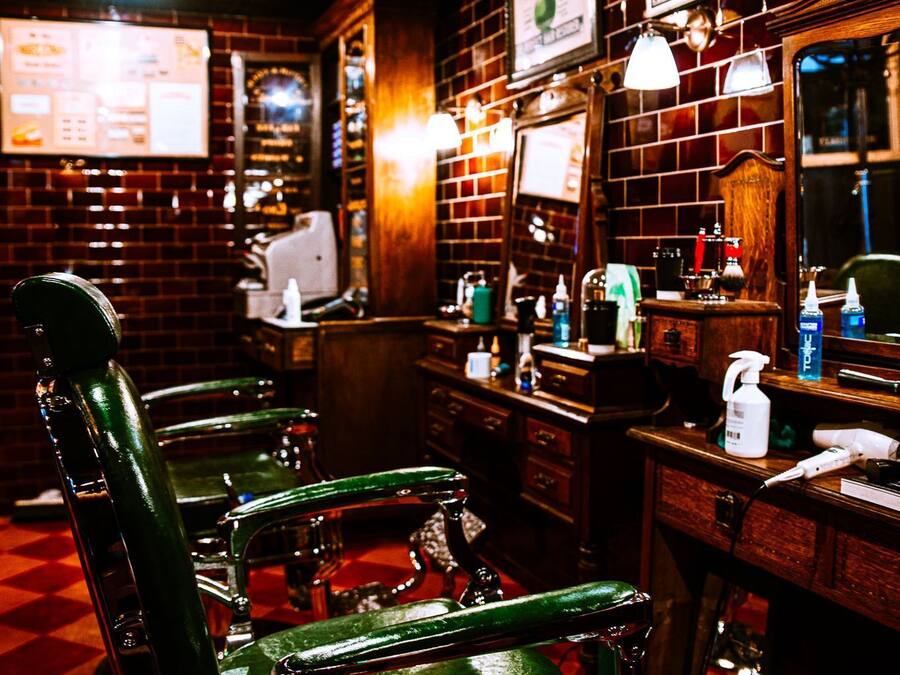 Una barbería con sillas de barbero verdes antiguas, una pared de ladrillo y carteles enmarcados.