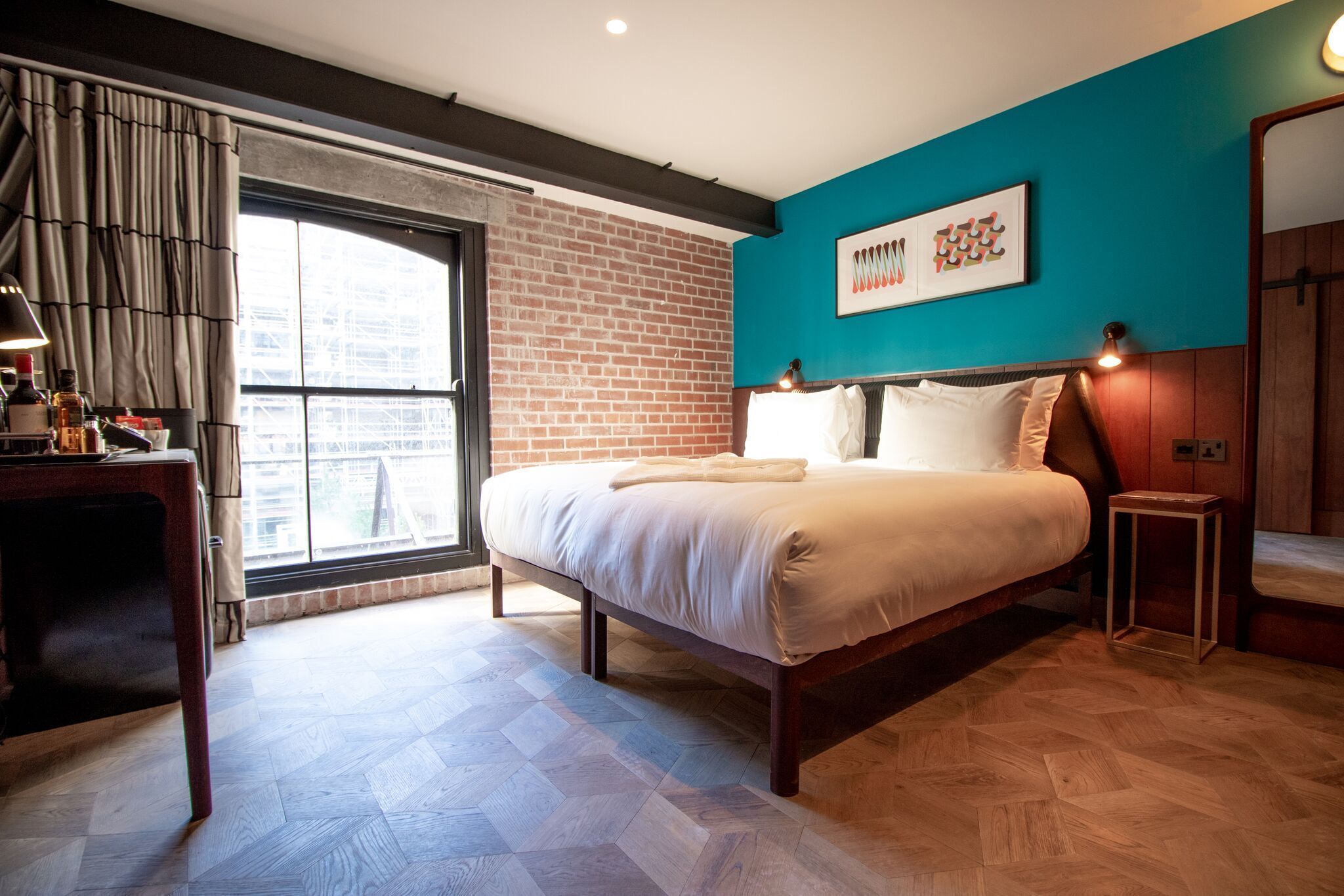 A modern hotel room with a large bed, a desk, and a view of the city through a large window.