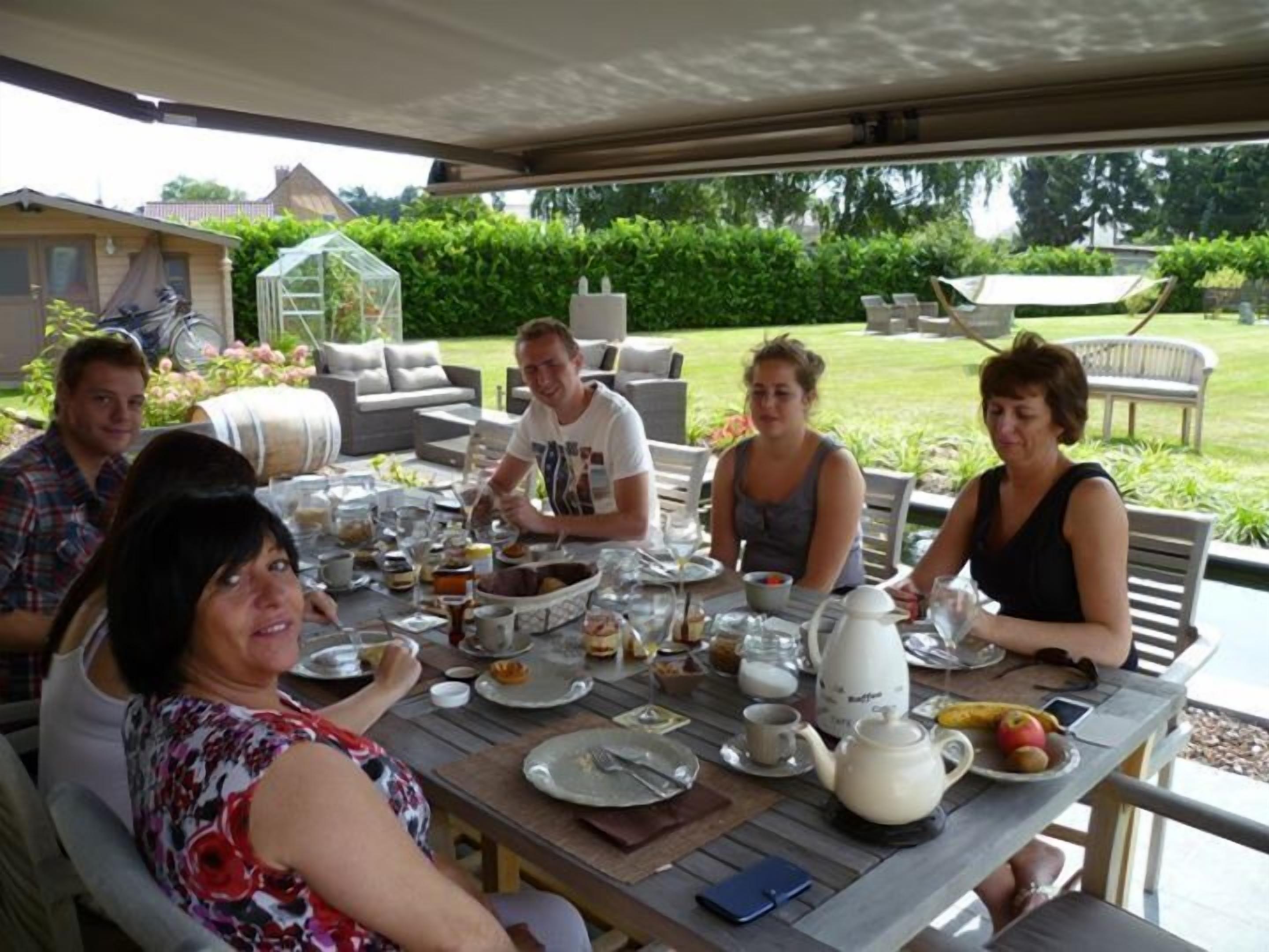 Un groupe de personnes est assis à une table en plein air, devant divers plats, une théière et, en arrière-plan, une serre.