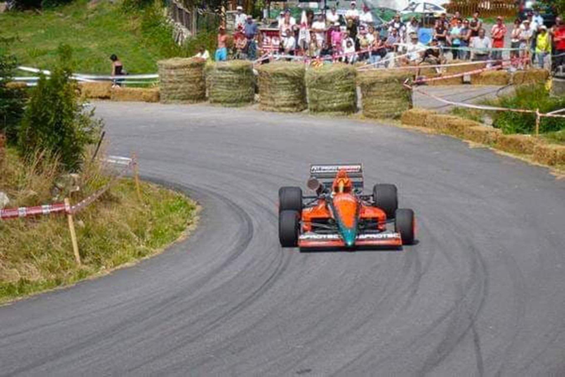 Un auto de carreras en una pista, con fardos de heno y espectadores de fondo.