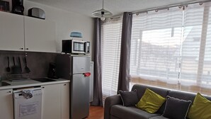 Un espacio habitable compacto con kitchenette, un sofá gris con cojines verde y negro, y una ventana con cortinas.