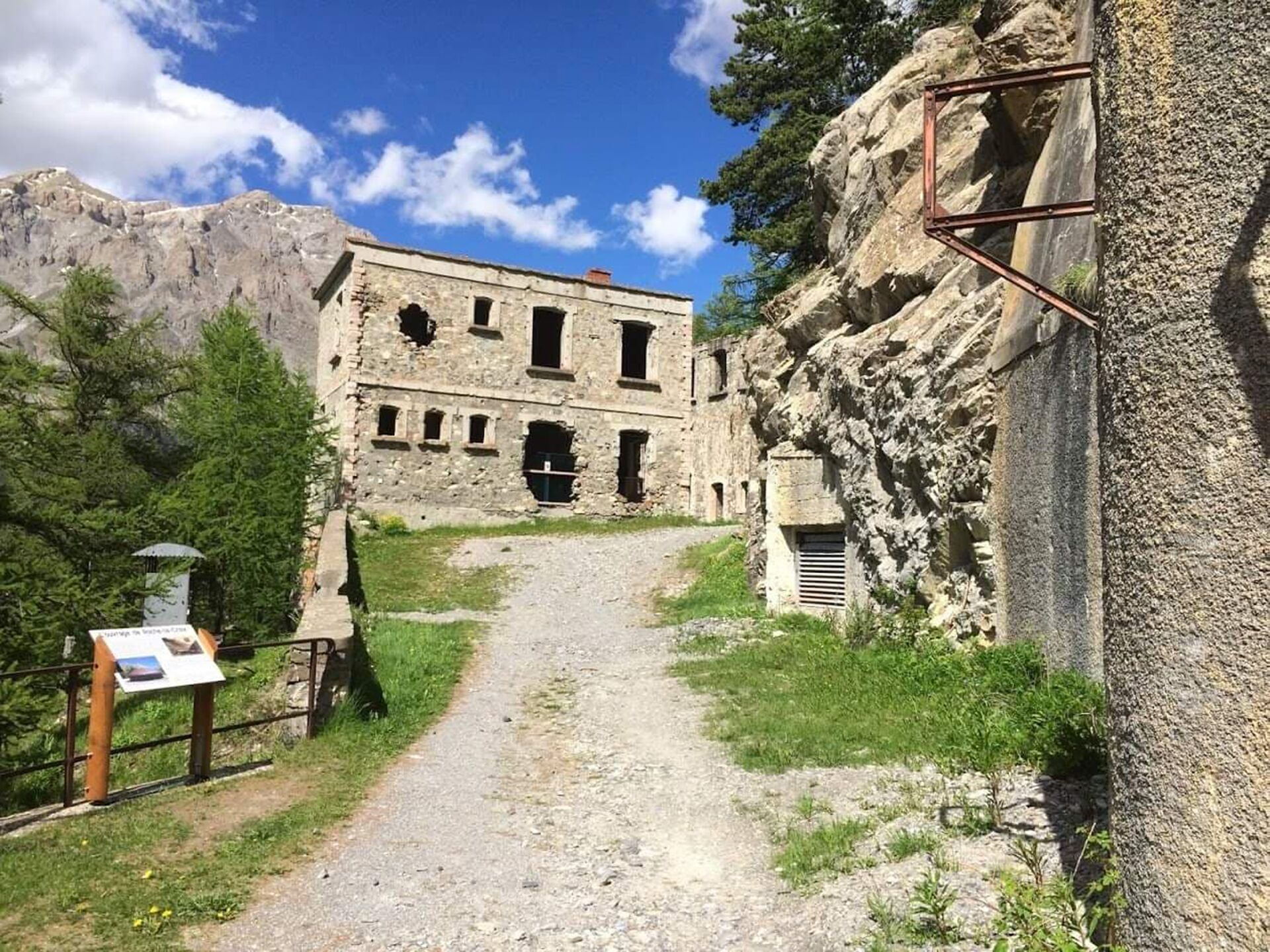 Un ancien bâtiment en pierre, auquel mène un sentier de terre, entouré de montagnes et de verdure.