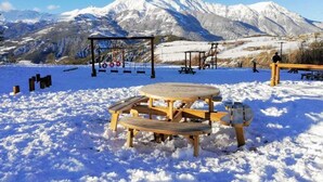 Un área de picnic cubierta de nieve con una mesa y bancos de madera, un columpio y vistas a montañas nevadas al fondo.