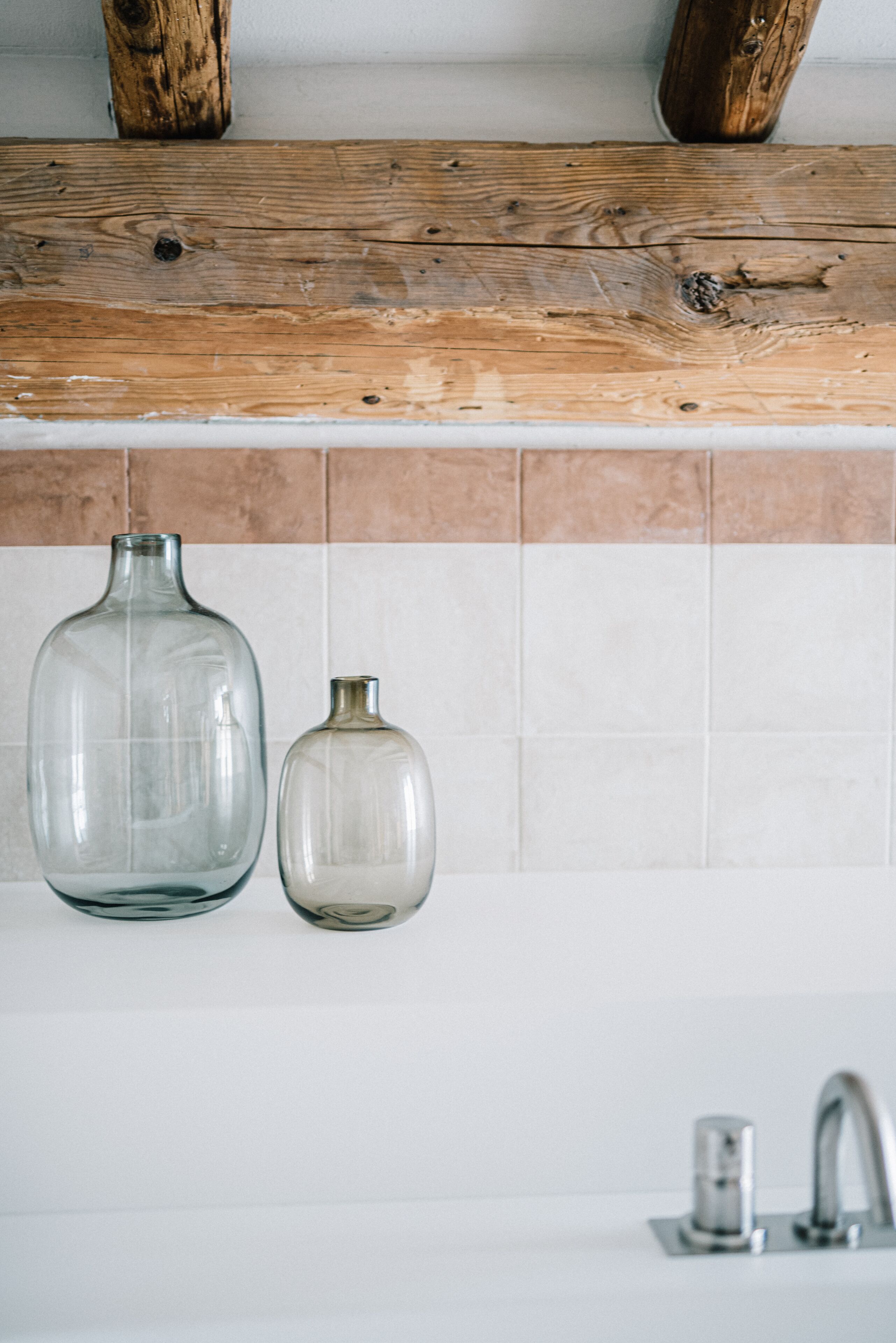 Une salle de bain avec une poutre en bois au-dessus d’un mur carrelé, un grand vase en verre transparent et un plus petit vase en verre transparent posé sur un lavabo blanc.