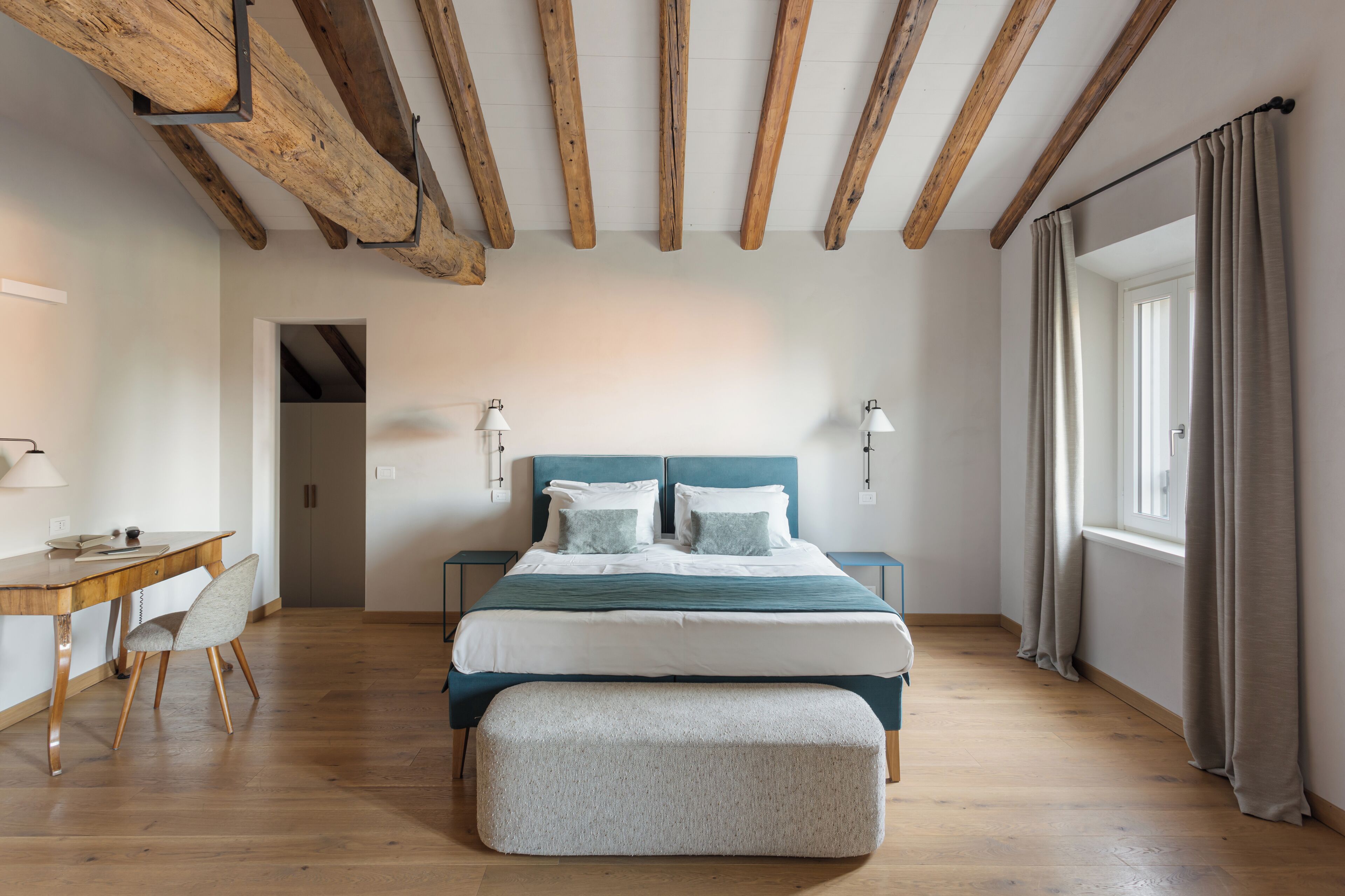 Ein Schlafzimmer mit Holzbalkendecke, einem Bett mit blauer Kopfteil, Nachttischlampen, einem Schreibtisch mit Stuhl und einem Fenster mit Vorhängen.