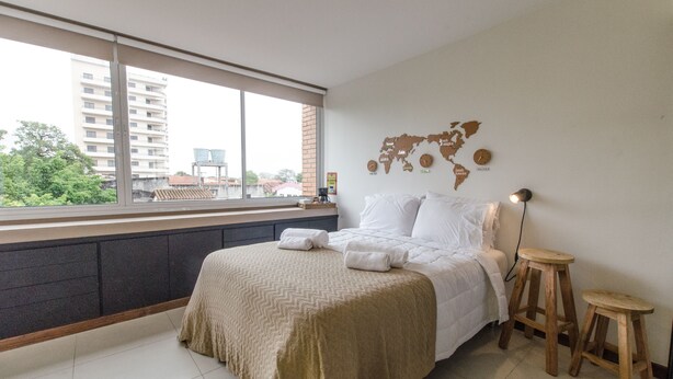 A hotel room with a large window, a bed with white linens, a world map on the wall, and two wooden stools. - Nuestra Sra. de la Asunción Stylish Loft (Asunción)