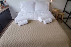 A neatly made bed with white linens and a beige patterned rug. There are two wooden stools beside the bed.