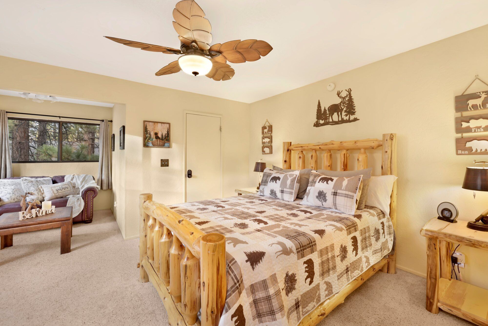 A bedroom with a wooden bed, a ceiling fan, a bedside table with a lamp, and a window with a view of trees.