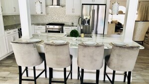 A modern kitchen with white cabinets, a stainless steel refrigerator, and a marble countertop. There are four chairs with a light fabric upholstery and black legs.