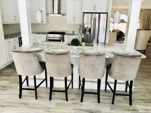 A modern kitchen with white cabinets, a stainless steel refrigerator, and a marble countertop. There are four chairs with a light fabric upholstery and black legs.