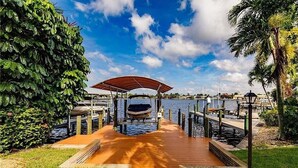 A wooden dock leading to a covered seating area by a body of water, surrounded by greenery and a clear sky.
