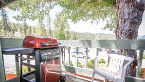 Ein überdachter Außenbereich mit Grill, einem weißen Plastikstuhl und Blick auf einen Parkplatz und Bäume.