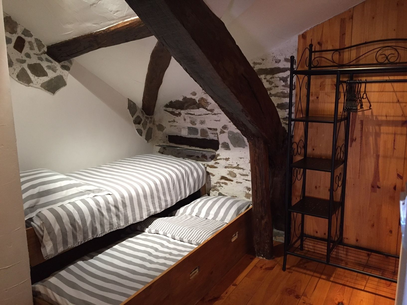 Une petite chambre rustique avec un lit simple, des poutres apparentes, un mur en pierre et une étagère en métal.