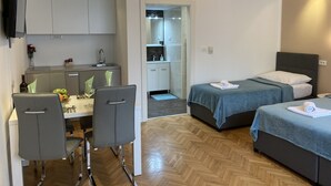 A modern hotel room with a kitchenette, two beds, a dining area with a table and chairs, and a TV mounted on the wall.