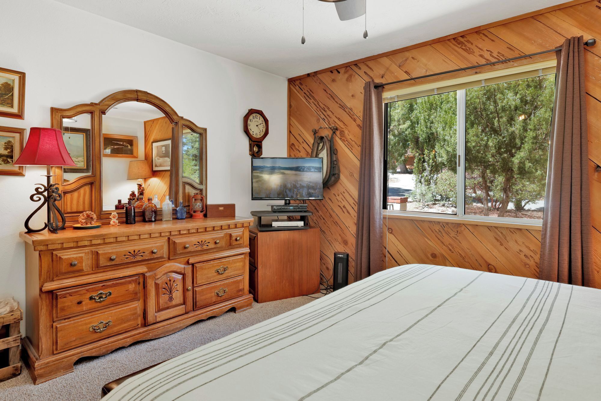 Un dormitorio con cabecera de madera, un ventanal con cortinas, un aparador de madera con cajones, un espejo, un televisor y una lámpara.