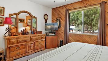 Un dormitorio con cabecera de madera, un ventanal con cortinas, un aparador de madera con cajones, un espejo, un televisor y una lámpara.