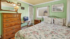 A bedroom with a wooden dresser, a bed with floral bedding, a desk with a computer, and a window with curtains.