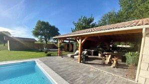 A swimming pool with a wooden deck and a covered patio area with outdoor furniture. - Le Refuge des Epicuriens (EYMET)
