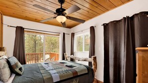 A bedroom with a bed, a ceiling fan, a window, and a view of trees. - Living Log Cabin#1494 (Big Bear City)