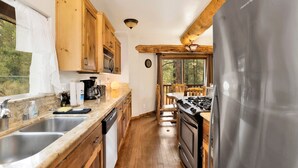 A kitchen with wooden cabinets, a stainless steel refrigerator, a stove, and a window overlooking a forest. - Living Log Cabin#1494 (Big Bear City)