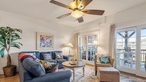 A living room with a blue sofa, a wicker chair, a coffee table, a ceiling fan, a potted plant, and a large window with curtains. - T's Retreat: Semi-oceanfront, third floor condo, two beach accesses close by. (Kill Devil Hills)