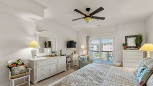 A bedroom with a bed, a dresser, a chair, a desk, a ceiling fan, a TV, and a balcony with a view of the city. - T's Retreat: Semi-oceanfront, third floor condo, two beach accesses close by. (Kill Devil Hills)