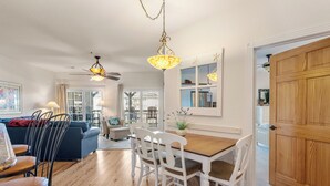 A spacious living room with a wooden dining table, white chairs, a blue sofa, and a ceiling fan. - T's Retreat: Semi-oceanfront, third floor condo, two beach accesses close by. (Kill Devil Hills)