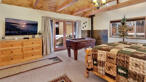 Een gezellige kamer met een houten plafond, een televisie, een tafelvoetbaltafel, een bed met een quilt en een raam met uitzicht op een besneeerd landschap.