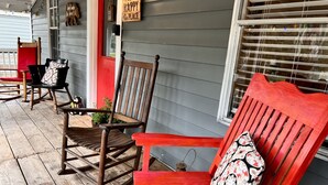 Une véranda avec des fauteuils à bascule en bois et un banc rouge, une enseigne en bois accrochée au mur et une fenêtre avec des stores.