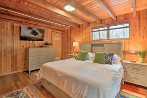 Ein Schlafzimmer mit Holzwänden, einem Bett, einem an der Wand montierten Fernseher, einem Fenster und einem Nachttisch mit Lampe.