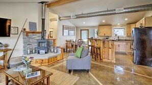 A spacious living area with a stone fireplace, a gray sofa, wooden furniture, and a kitchen with a black refrigerator. - 'Coastal Soul' Arcata Area California Home (Blue Lake)