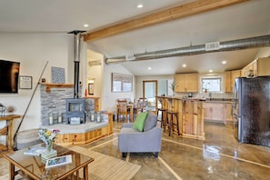 A spacious living area with a stone fireplace, a gray sofa, wooden furniture, and a kitchen with a black refrigerator.