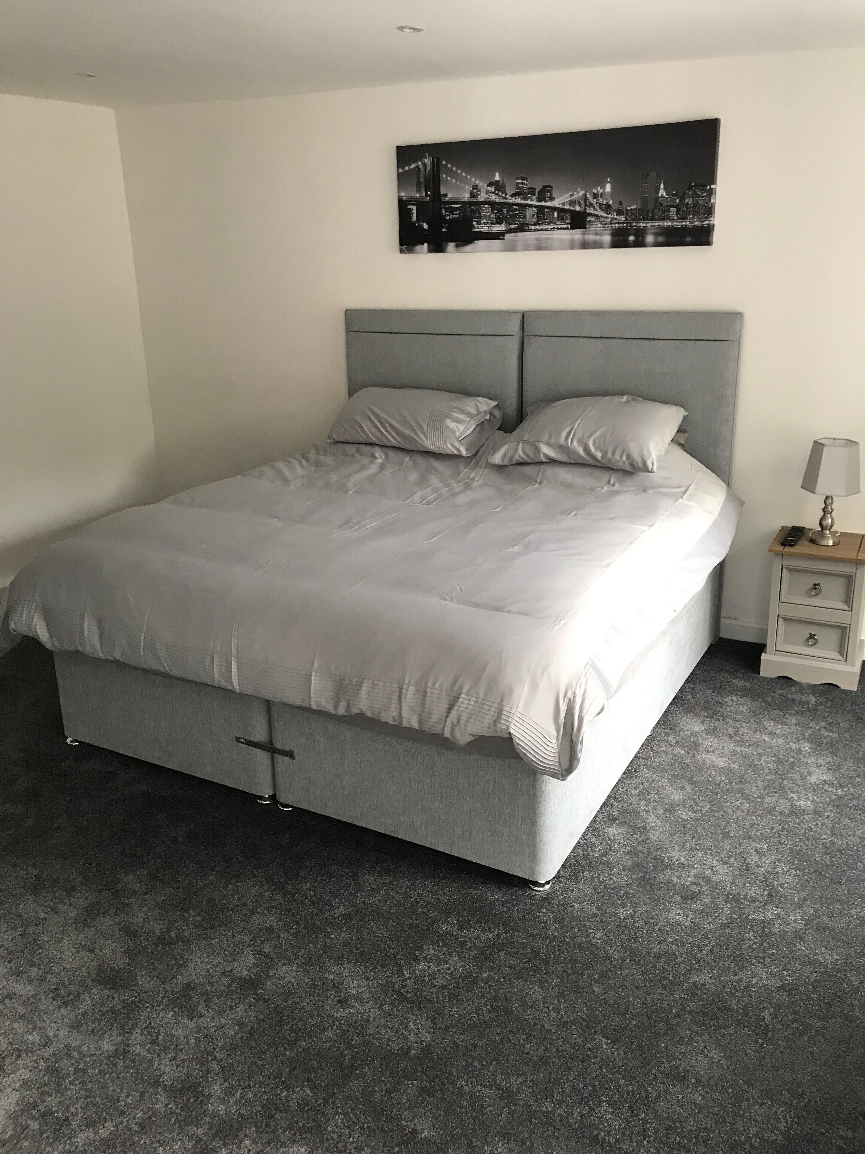 Un letto ben rifatto con una testiera grigia e cuscini, un comodino con una lampada e un quadro con una veduta della città.