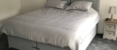 Un letto ben rifatto con una testiera grigia e cuscini, un comodino con una lampada e un quadro con una veduta della città.