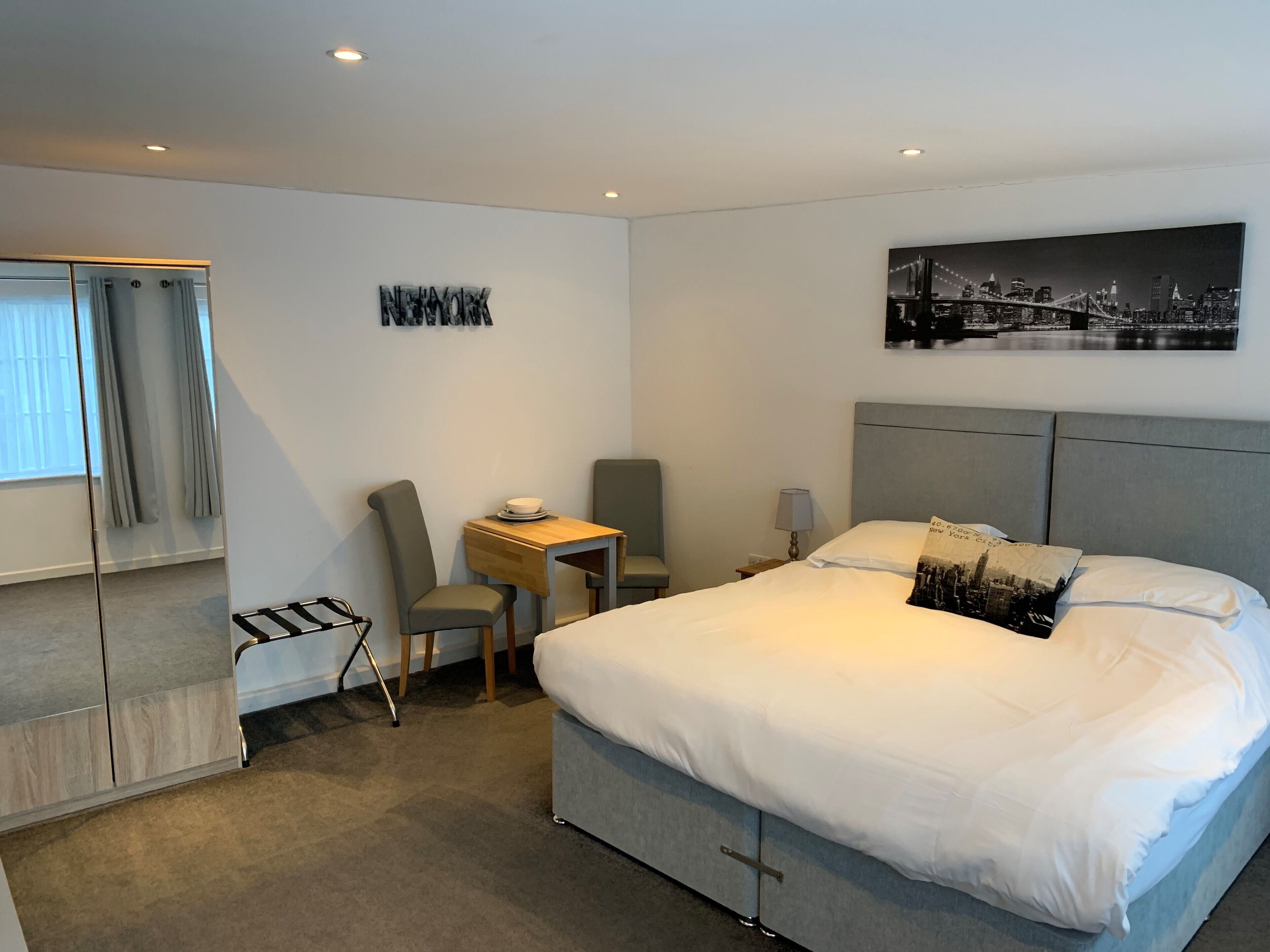 Ett modernt hotellrum med en stor säng, ett skrivbord, en stol, en garderob och ett konstverk med en stadsbild på väggen.