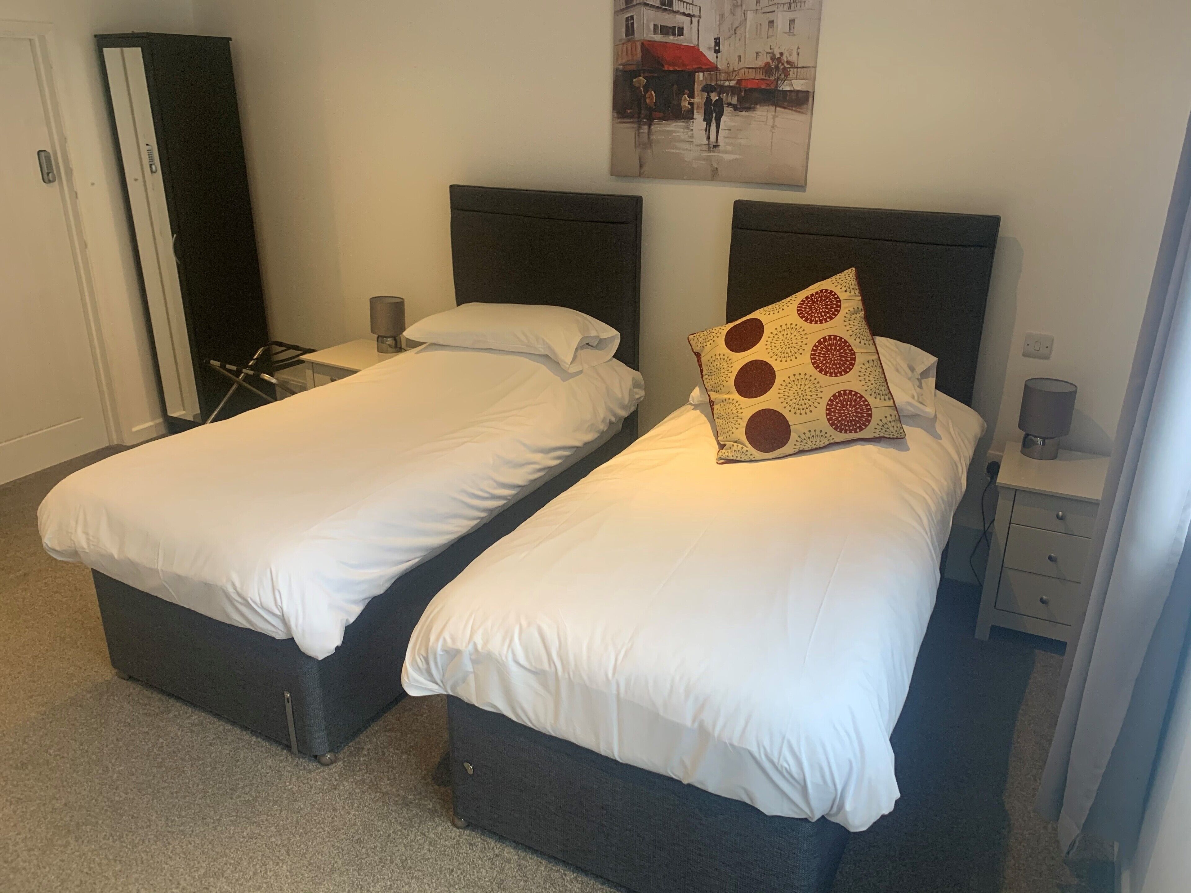 Habitación doble con dos camas individuales, una mesita de noche con lámpara, un cuadro en la pared y una puerta.