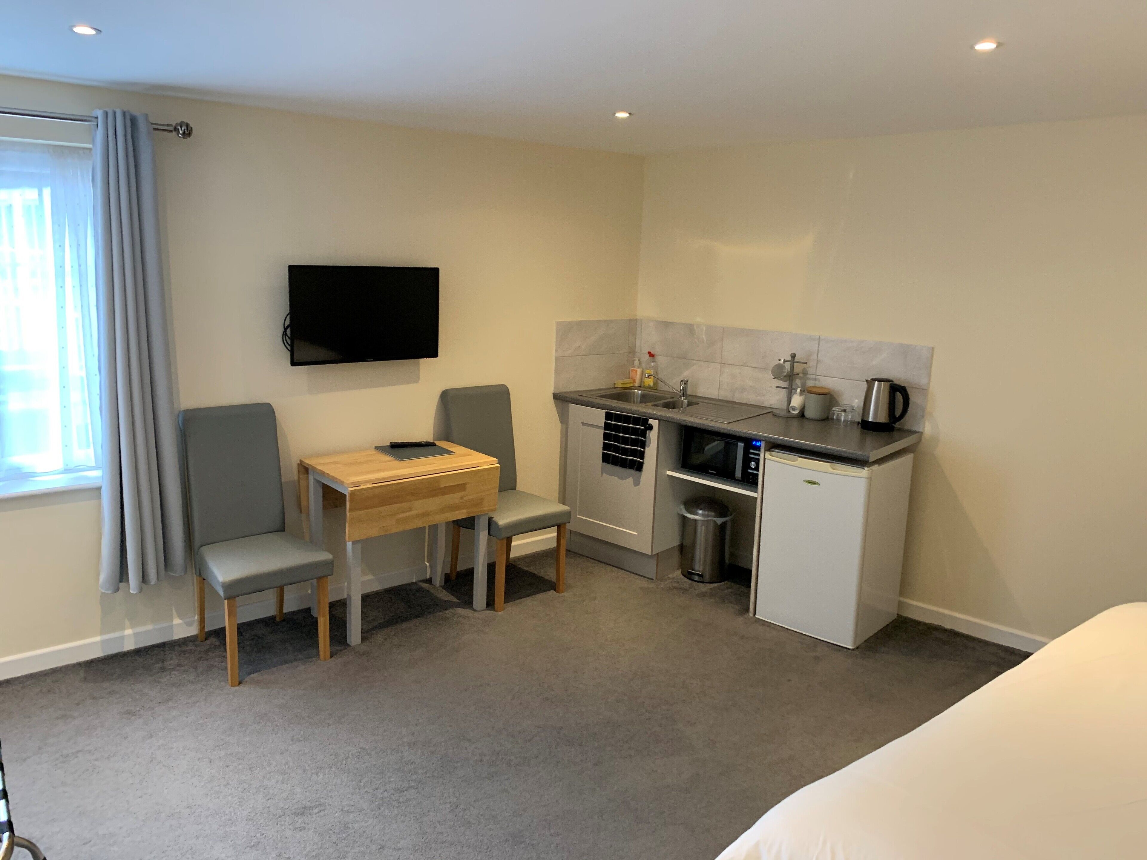 A compact hotel room with a kitchenette, a flat-screen TV mounted on the wall, a small dining table with two chairs, and a single bed.