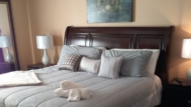 A neatly made bed with a headboard, a bedside table with a lamp, and a painting on the wall. - Entertainment 4539 (Snellville)
