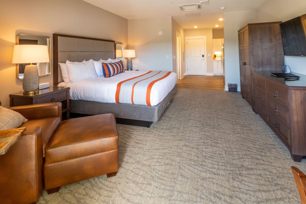A hotel room with a large bed, a wooden dresser, a desk with a lamp, and a leather armchair.