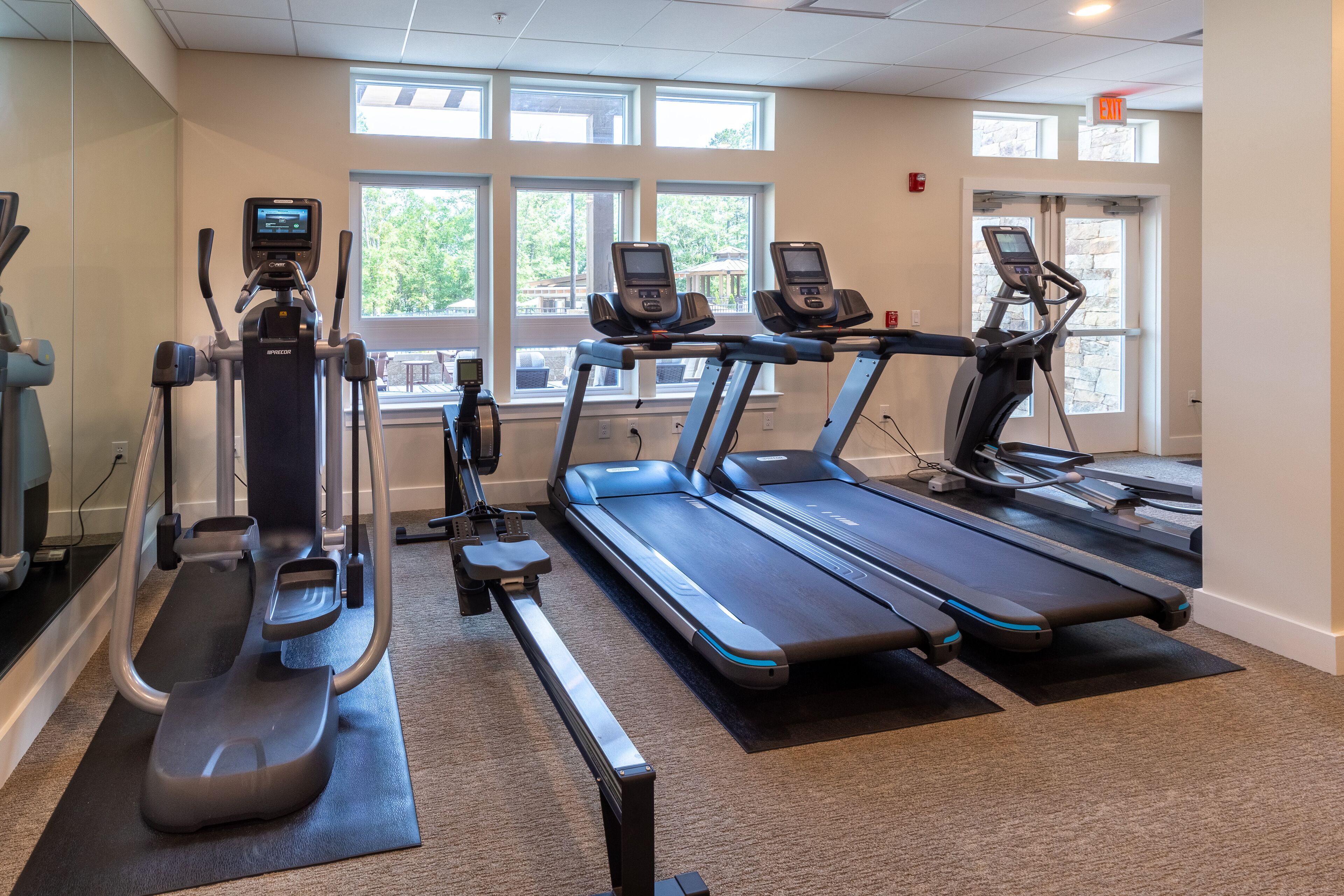 A gym with treadmills, an elliptical, and a stationary bike, with large windows and an exit sign.