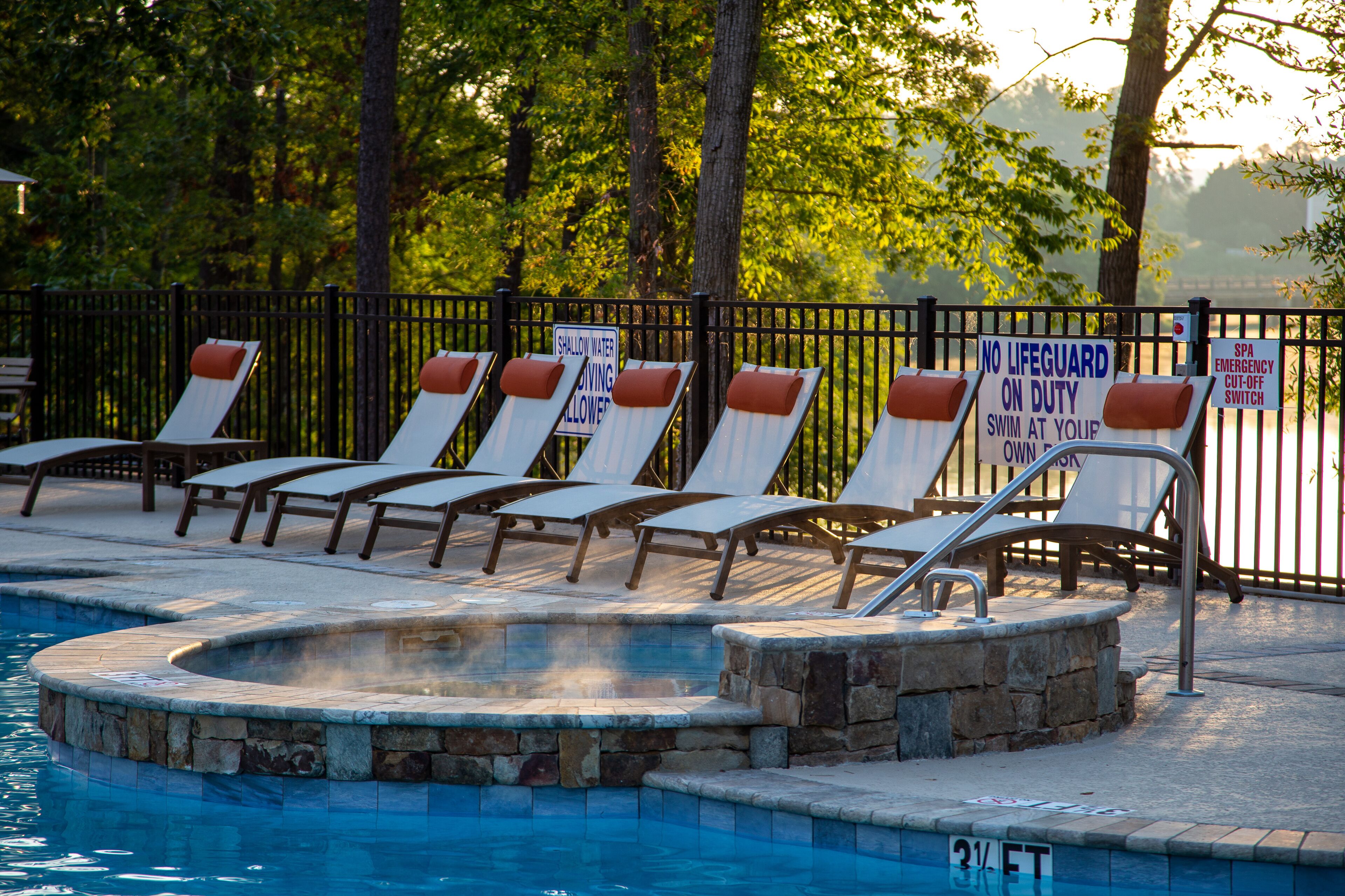 A pool area with lounge chairs and a hot tub, surrounded by a fence and trees.
